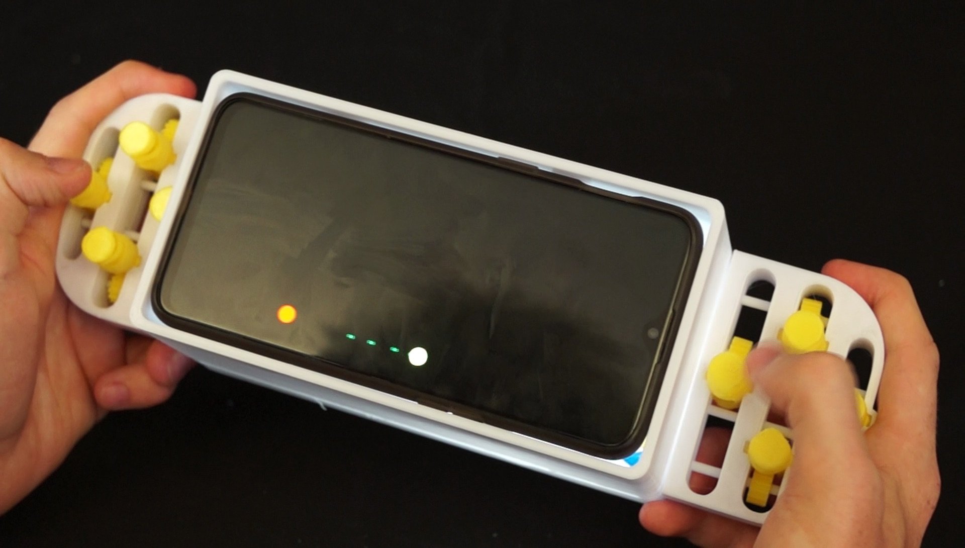 Hands hold a game controller case with a smartphone inserted. The controller
has yellow knobs and buttons, and the phone screen shows small colorful
lights.
